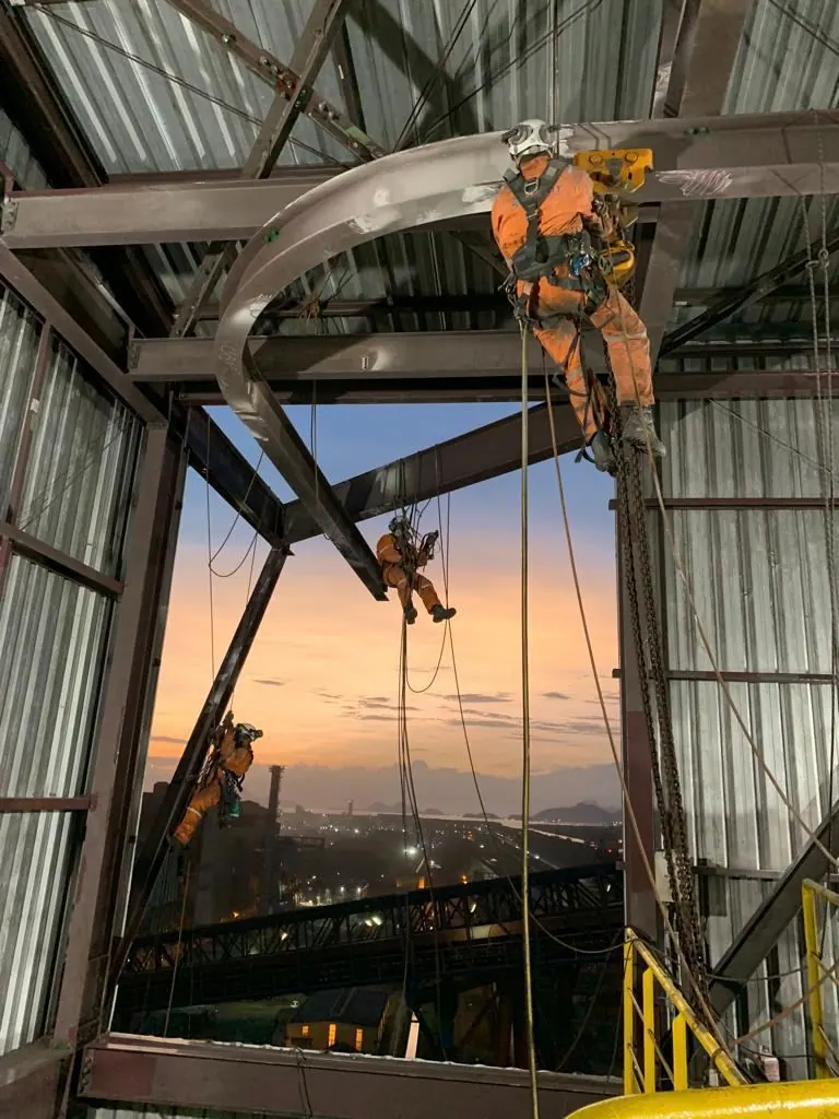 Operadores em rapel no arco estrutural de um galpão ao entardecer